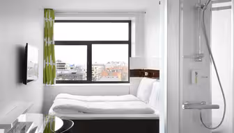 Kompakt moderne hotelværelse med et stort vindue, der viser et bybillede. Værelset har en seng med hvidt sengetøj, et vægmonteret tv, et skrivebord med glasplade med en stol og et brusebad med glasdøre. Lys og minimalistisk indretning med grønne mønstrede gardiner.