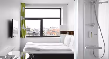 Kompakt moderne hotelværelse med et stort vindue, der viser et bybillede. Værelset har en seng med hvidt sengetøj, et vægmonteret tv, et skrivebord med glasplade med en stol og et brusebad med glasdøre. Lys og minimalistisk indretning med grønne mønstrede gardiner.