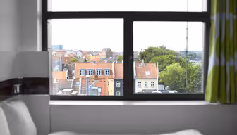 Udsigt gennem et vindue, der viser et bybillede med bygninger med røde tag og grønt. Værelsesindretningen har en seng med hvidt sengetøj og et gardin med grønne og hvide mønstre. Himlen er overskyet.