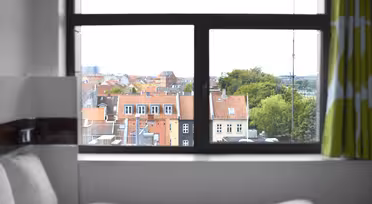 Udsigt gennem et vindue, der viser et bybillede med bygninger med røde tag og grønt. Værelsesindretningen har en seng med hvidt sengetøj og et gardin med grønne og hvide mønstre. Himlen er overskyet.