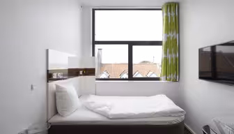 Moderne, minimalistisk soveværelse med en seng ved siden af et stort vindue. Vinduet har grønne og hvide gardiner og giver udsigt til hustage. Et fladskærms-tv er monteret på væggen, og der er et lille, rundt glas sidebord ved sengen.