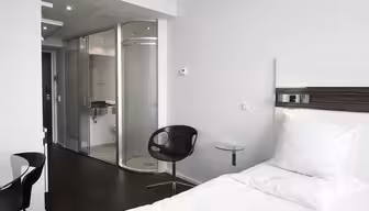Et moderne hotelværelse med en pænt lavet enkeltseng, en rund sort stol og et sidebord med glasplade. Værelset har et glasbadeværelse med bruser, elegant mørkt trægulv og en minimalistisk indretning.