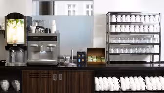 En kaffestation med en kaffemaskine, en dispenser til varme drikke, stablede kopper, glas og en trækasse med teposer. Hvide krus er arrangeret på hylder og under disken. Baggrunden viser et matteret glaspanel og et vindue.