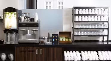 En kaffestation med en kaffemaskine, en dispenser til varme drikke, stablede kopper, glas og en trækasse med teposer. Hvide krus er arrangeret på hylder og under disken. Baggrunden viser et matteret glaspanel og et vindue.