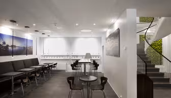 Moderne cafeinteriør med sorte siddepladser og små træborde til venstre. Hvide vægge viser kunst med maritimt tema. I midten en disk med barstole. Vindeltrappe til højre, der fører op med planter synlige i baggrunden.