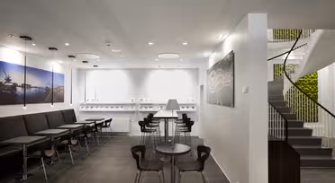 Moderne cafeinteriør med sorte siddepladser og små træborde til venstre. Hvide vægge viser kunst med maritimt tema. I midten en disk med barstole. Vindeltrappe til højre, der fører op med planter synlige i baggrunden.