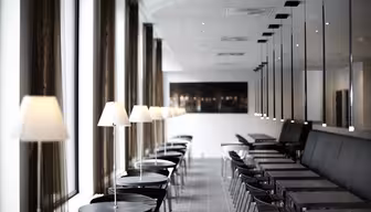 Et moderne, minimalistisk cafeinteriør med rækker af sorte borde og stole, foret med hvide lamper. Store vinduer til venstre lukker naturligt lys ind, mens spejle til højre skaber reflekser, der forstærker den rummelige fornemmelse.