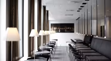 Et moderne, minimalistisk cafeinteriør med rækker af sorte borde og stole, foret med hvide lamper. Store vinduer til venstre lukker naturligt lys ind, mens spejle til højre skaber reflekser, der forstærker den rummelige fornemmelse.