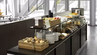 En moderne buffetopstilling med en række forskellige fødevarer, herunder rundstykker, frugt, korn og juice. Tallerkener og redskaber er pænt organiseret. Serveringsområdet har et slankt, moderne design med masser af naturligt lys.
