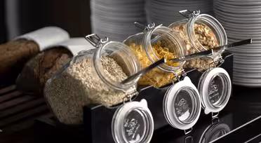 Tre glaskrukker fyldt med forskellige kornsorter - havre, cornflakes og granola - er åbne på et sort stativ, hver med en metalske. I baggrunden er der stablet hvide tallerkener og nogle skiver brød på et skærebræt.