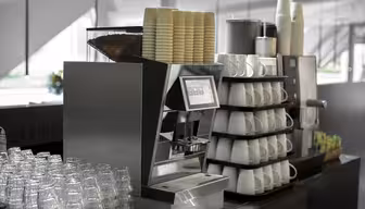 En elegant, moderne kaffemaskine på en disk, omgivet af stakke af papirkopper, keramiske krus og glaskopper. Rammerne ser ud til at være en café eller et pauseområde med stærkt, naturligt lys.