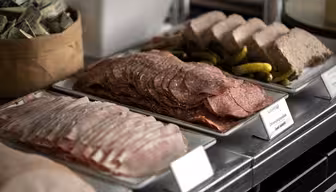 En buffetudstilling med bakker med diverse delikatessekød, inklusive skiver af salami og skinke, sammen med syltede garniture. En kurv fyldt med pakket smør er synlig i baggrunden.