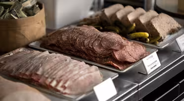 En buffetudstilling med bakker med diverse delikatessekød, inklusive skiver af salami og skinke, sammen med syltede garniture. En kurv fyldt med pakket smør er synlig i baggrunden.