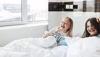 To børn griner i hvidt sengetøj nær et vindue. Barnet til venstre har lyst hår og er iført en hvid skjorte med farverige bogstaver. Barnet til højre har brunt hår. Begge sidder op og smiler glad.