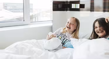 To børn griner i hvidt sengetøj nær et vindue. Barnet til venstre har lyst hår og er iført en hvid skjorte med farverige bogstaver. Barnet til højre har brunt hår. Begge sidder op og smiler glad.
