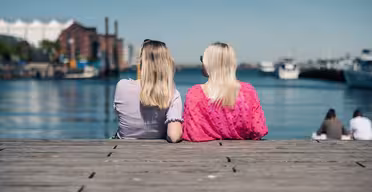 To kvinder med lyst hår sidder på en trædok, med front mod en rolig vandmasse. Den ene har en lyserød skjorte på, den anden en lyslilla. Et par både er synlige på vandet, med bygninger i baggrunden under en klar blå himmel.