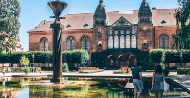 Et par går nær et rektangulært springvand med en skulptur foran en historisk murstensbygning med buede vinduer og et symmetrisk design. Scenen er sat i en solrig have med klippede hække og blomsterbede.