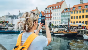 En person med en gul rygsæk tager et billede af farverige bygninger langs en kanal med både og fodgængere i nærheden. Scenen fanger et livligt, livligt område ved havnefronten under en overskyet himmel.