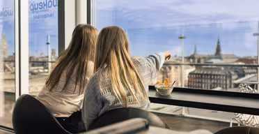 To personer sad indendørs ved et vindue, den ene peger ud mod et bybillede med hustage og spir. En skål med snacks står på bordet. Scenen antyder en afslappet atmosfære med panoramaudsigt.