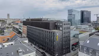 Luftfoto af et bybillede med en moderne, mørkfarvet hotelbygning med navnet "Wakeup" på siden. De omkringliggende bygninger omfatter ældre bygninger med gråt tage, moderne glasbygninger og delvist overskyet himmel.