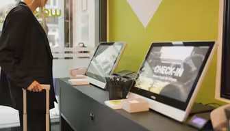 En person står ved en check-in skranke på hotellet og bruger selvbetjeningskiosker. Kioskerne viser ordet CHECK-IN på deres skærme. Værelset har et moderne design med limegrønne og sorte accenter. Personen holder et kufferthåndtag.