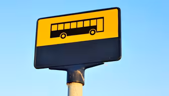 Gult og sort busstoppestedsskilt med et busikon, monteret på en stang mod en klar blå himmel.