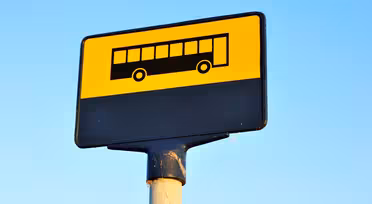 Gult og sort busstoppestedsskilt med et busikon, monteret på en stang mod en klar blå himmel.