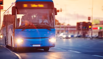 En blå bus kører ned ad en gade ved solnedgang. Bussens lys er tændt, og destinationsskiltet lyser. Vejen og det omkringliggende område er let sløret, hvilket indikerer bevægelse. Biler og andre byelementer er synlige i baggrunden.