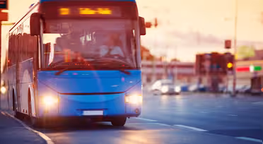 En blå bus kører ned ad en gade ved solnedgang. Bussens lys er tændt, og destinationsskiltet lyser. Vejen og det omkringliggende område er let sløret, hvilket indikerer bevægelse. Biler og andre byelementer er synlige i baggrunden.