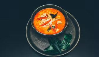 En skål med livlig rød karrysuppe pyntet med friske krydderurter og skiver rød chili står på en mørk tallerken. Ved siden af skålen er kaffirlimeblade arrangeret på tallerkenen, hvilket skaber en frisk og appetitlig præsentation.