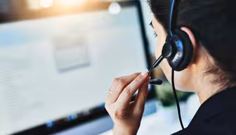 En kvinde iført headset sidder ved et skrivebord og fokuserer på en computerskærm. Sollys oplyser blidt hendes arbejdsområde.