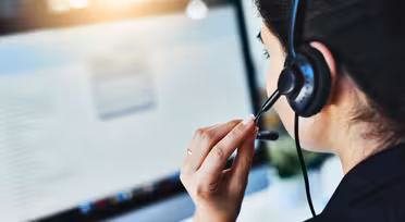 En kvinde iført headset sidder ved et skrivebord og fokuserer på en computerskærm. Sollys oplyser blidt hendes arbejdsområde.