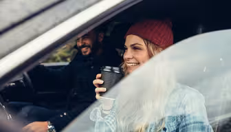 En kvinde i en rød hue sidder i en bil, holder en kaffekop og smiler. Hun sidder på passagersædet ved siden af en mand, der kører og smiler, begge i afslappet påklædning. Scenen er fanget gennem det åbne bilvindue.