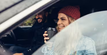 En kvinde i en rød hue sidder i en bil, holder en kaffekop og smiler. Hun sidder på passagersædet ved siden af en mand, der kører og smiler, begge i afslappet påklædning. Scenen er fanget gennem det åbne bilvindue.