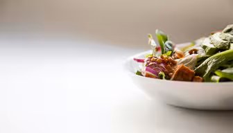 En hvid skål fyldt med en frisk salat ses med grønt, rødløg, linser og croutoner. Fadet stilles på en lys overflade med en blød, neutral baggrund.