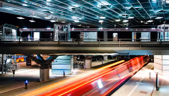 Lang eksponeringsfoto af en busstation om natten. Striber af rødt og orange lys antyder et køretøj i bevægelse under en oplyst, moderne glas- og metalstruktur. Cykler og en parkeret bus er synlige i baggrunden.