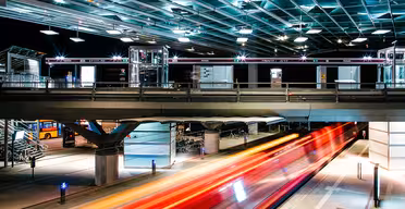 Lang eksponeringsfoto af en busstation om natten. Striber af rødt og orange lys antyder et køretøj i bevægelse under en oplyst, moderne glas- og metalstruktur. Cykler og en parkeret bus er synlige i baggrunden.