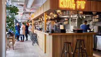 Et moderne, hyggeligt restaurantinteriør med trædisk og barstole. På neonskiltet står "KUNG FU." Flere personer står i nærheden af baren, og en medarbejder arbejder bag disken. Varm belysning og grønne planter tilføjer stemningen.