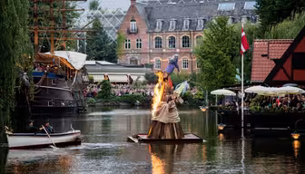 En træskulptur i brand flyder på en sø omgivet af tilskuere. Bygninger med store vinduer er i baggrunden. En person i en robåd nærmer sig den brændende skulptur. Et dansk flag er synligt nær en restaurant med udendørs siddepladser.