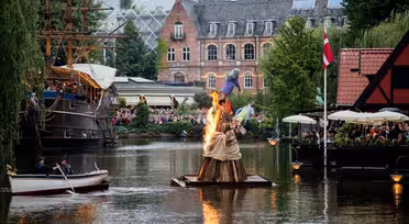 En træskulptur i brand flyder på en sø omgivet af tilskuere. Bygninger med store vinduer er i baggrunden. En person i en robåd nærmer sig den brændende skulptur. Et dansk flag er synligt nær en restaurant med udendørs siddepladser.