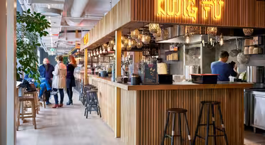 Et moderne restaurantinteriør med trædisk og barstole. Et neonskilt ovenfor læser "Kung Fu." Lånere ses interagere i baggrunden, og køkkenområdet er synligt bag disken. Planter og dekorative lys pryder rummet.