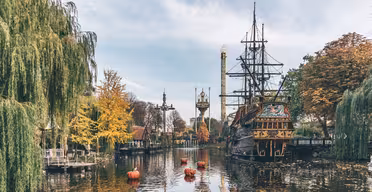 En rolig søscene med et forankret piratskib, omgivet af efterårstræer med gule og orange blade. Røde bøjer flyder i vandet, og et eventyrslot er synligt i baggrunden under en overskyet blå himmel.