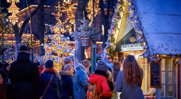 Folk samles til et festligt udendørsmarked om aftenen med en snedækket bod og farverige lys. En kvinde i rød frakke står ved boden. Dekorative hængelamper og oplyste træer skaber en varm, livlig atmosfære.