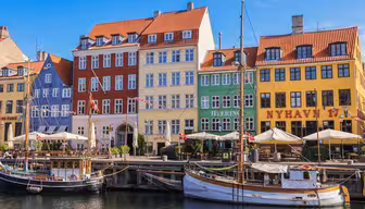 Naturskøn udsigt over Nyhavn, København, med farverige bygninger med røde, blå, grønne og gule facader. Træbåde lægger til langs vandet under en klar blå himmel. Patio paraplyer og mennesker er synlige nær havnefronten.