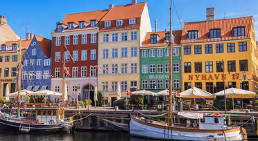 Naturskøn udsigt over Nyhavn, København, med farverige bygninger med røde, blå, grønne og gule facader. Træbåde lægger til langs vandet under en klar blå himmel. Patio paraplyer og mennesker er synlige nær havnefronten.