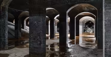 Et svagt oplyst underjordisk vandreservoir med buede murstenssøjler, der kaster skygger og refleksioner på det fugtige betongulv. Arkitekturen skaber en symmetrisk og uhyggelig atmosfære.