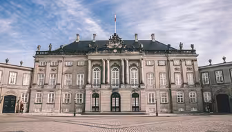 Elegant historisk bygning med en symmetrisk facade med udsmykkede vinduer og statuer på taget. Strukturen er en del af et større kompleks, sat mod en delvist overskyet himmel. Jorden er brostensbelagt, hvilket tilføjer den klassiske æstetik.