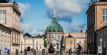 To personer cykler på en bred gade flankeret af historiske bygninger i København. Den grønkuplede Frederiks Kirke ses i baggrunden under en delvist overskyet himmel.