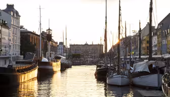 Udsigt til solnedgang af en kanal med forankrede sejlbåde på begge sider. Historiske bygninger ligger langs havnefronten og afspejler sig i det rolige vand. Himlen er blødt oplyst i varme nuancer, hvilket skaber en fredfyldt atmosfære.