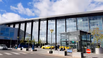 Udvendig udsigt af en moderne lufthavnsterminal med en glasfacade. Flere gule taxaer og et par personer står foran indgangen. Himlen er delvist overskyet, og der er træer og gadeskilte i nærheden af terminalen.